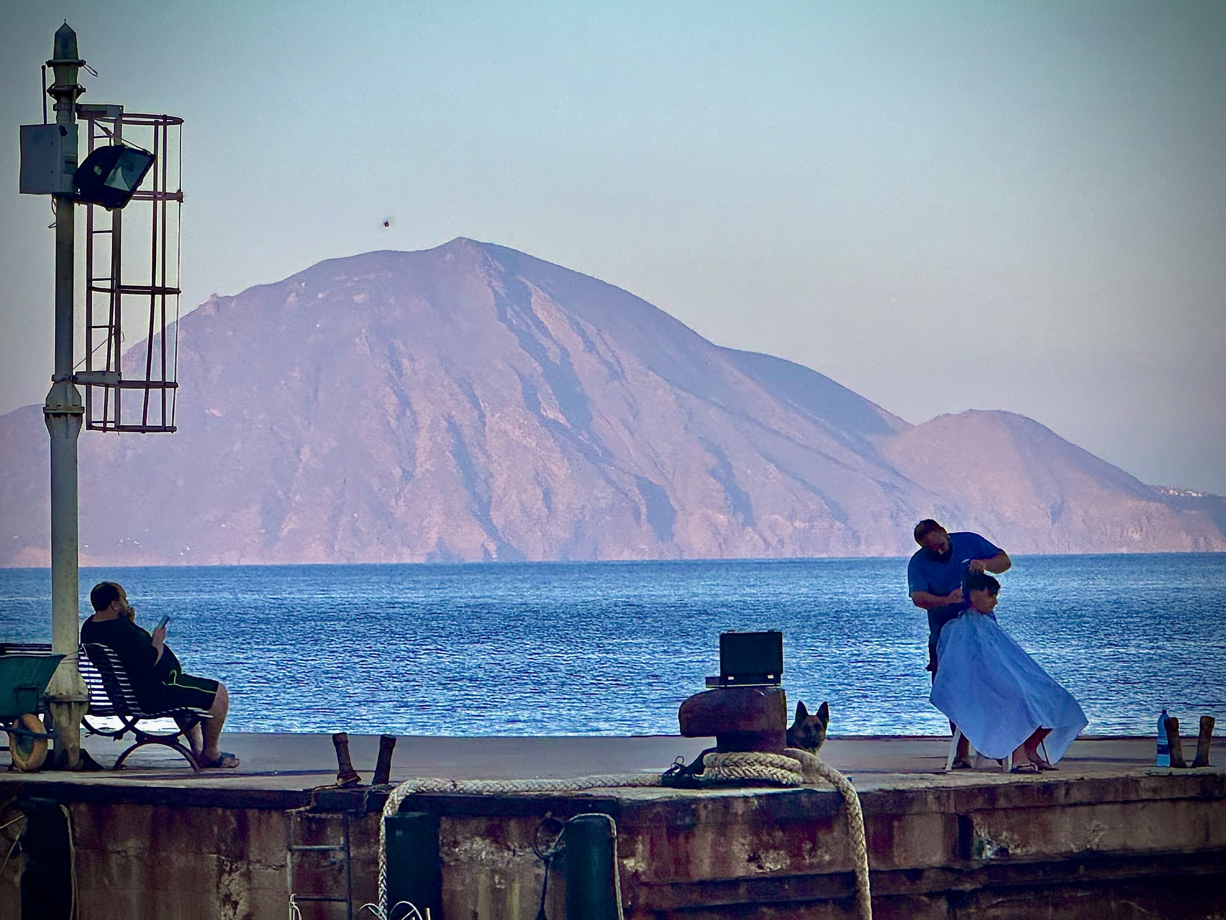 Ein Friseur rasiert ein Kind am Ende des Stegs von Alicudi, Filicudi im Hintergrund